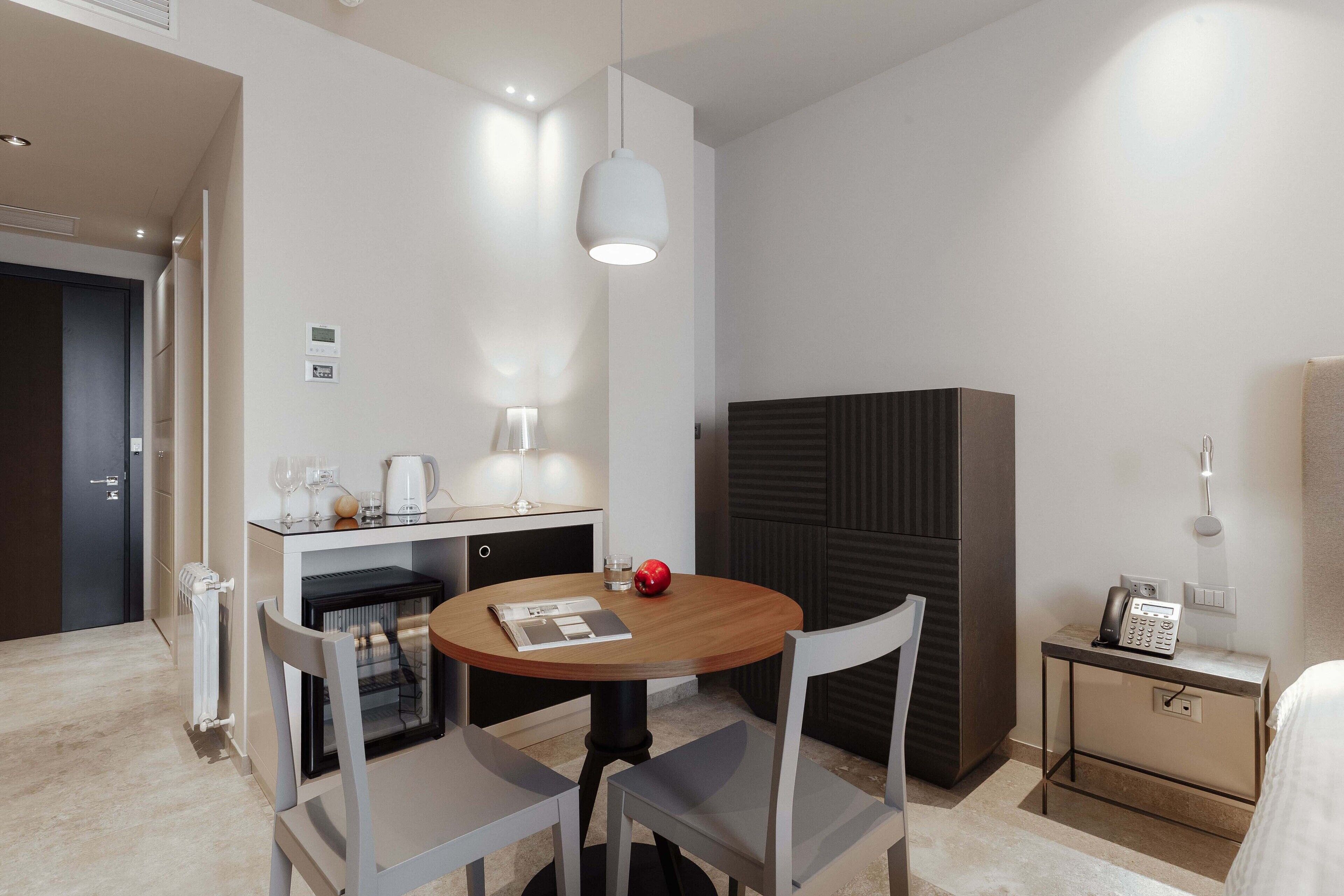 A modern hotel room with a round dining table, two chairs, a small kitchen area with a refrigerator, a coffee maker, and a telephone.