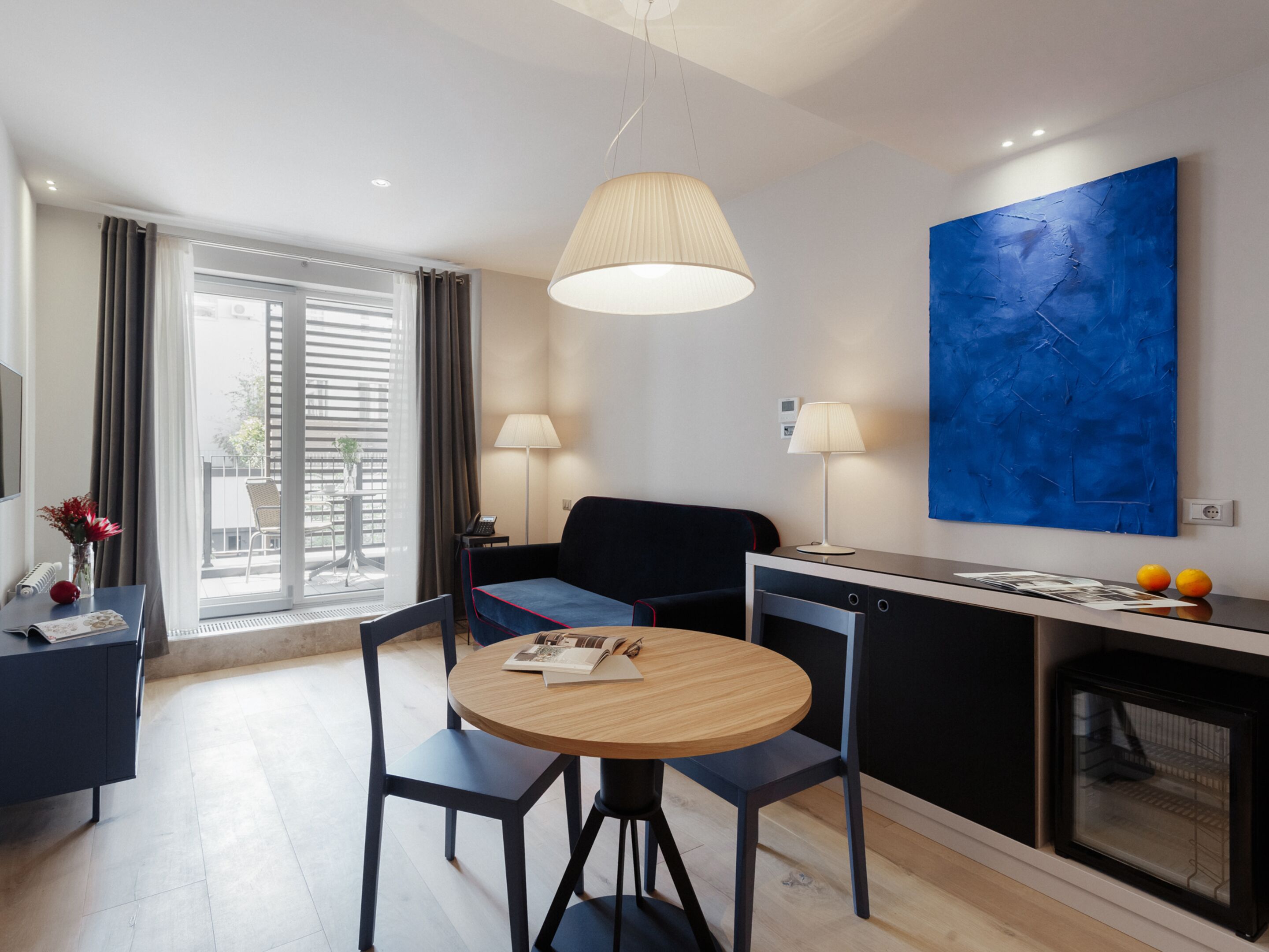 A modern hotel room with a dining area, a sofa, a round table, and a large blue painting on the wall.
