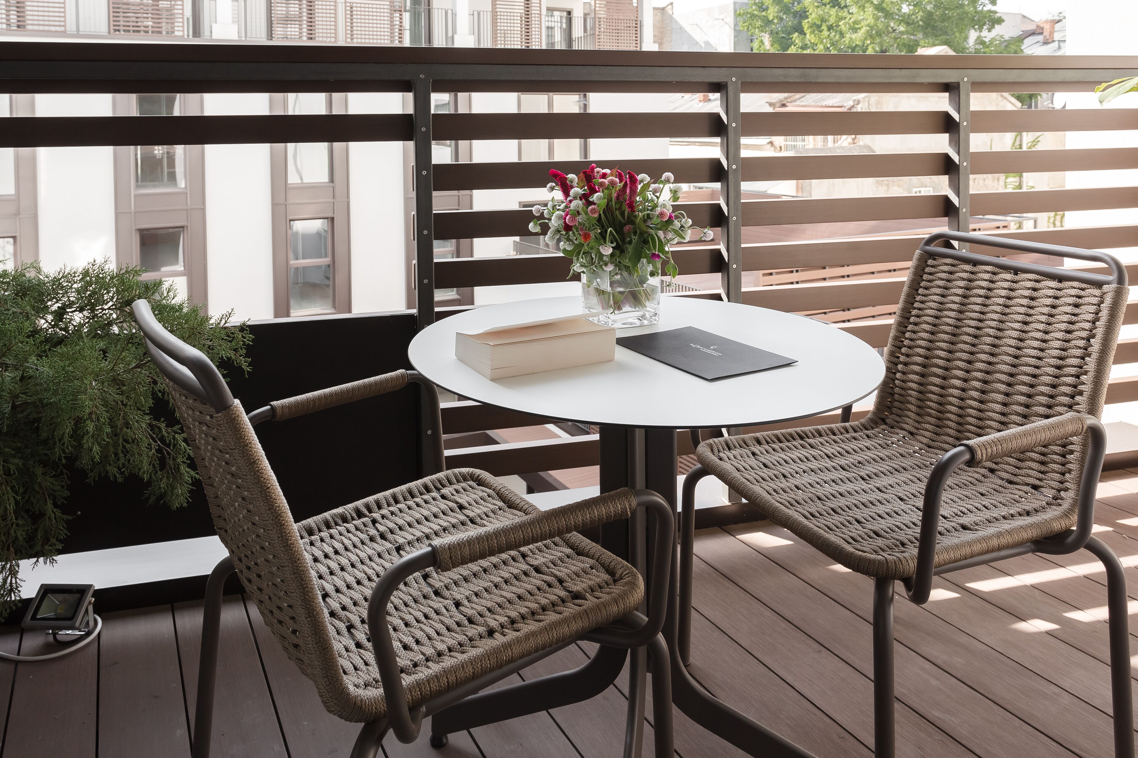 A balcony with wicker chairs, a round table with a book and a vase of flowers, and a view of buildings.