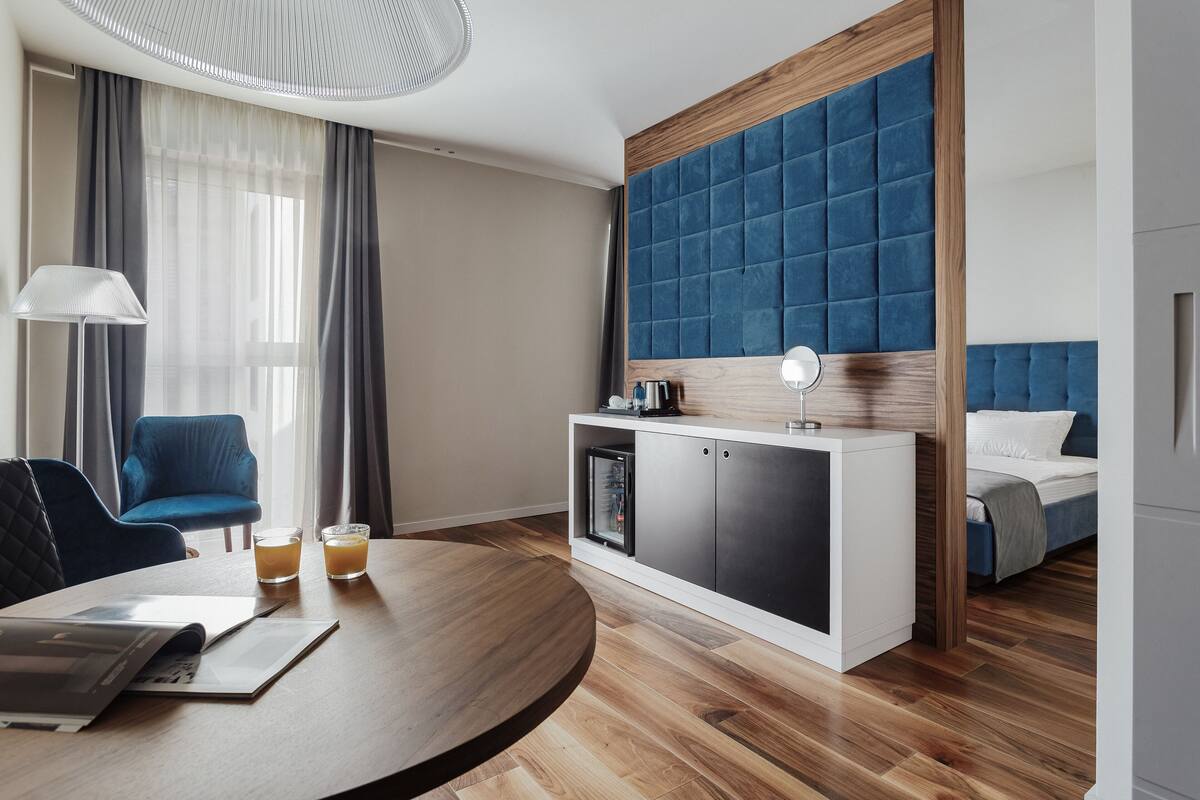 A modern hotel room with a round wooden table, a blue armchair, a bed with a blue headboard, and a minibar.