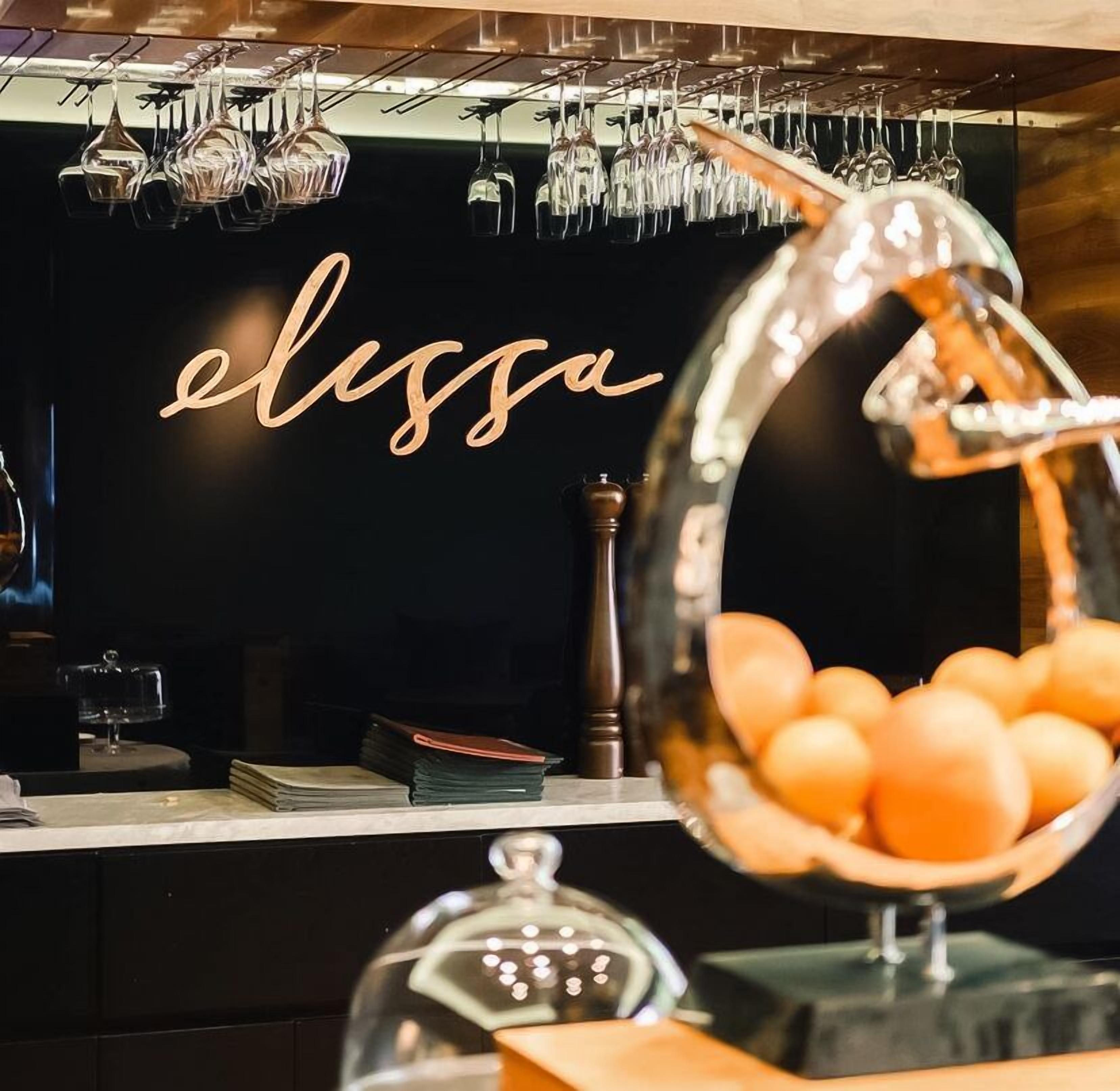 Un coin bar avec un mur noir et une enseigne sur laquelle on peut lire « elissa ». Des verres à vin sont accrochés au mur et un bol rempli d’oranges est posé sur le comptoir.