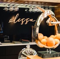 Una zona de bar con una pared negra y un letrero que dice "elissa". Hay copas de vino colgadas en la pared y un cuenco de naranjas sobre la barra.