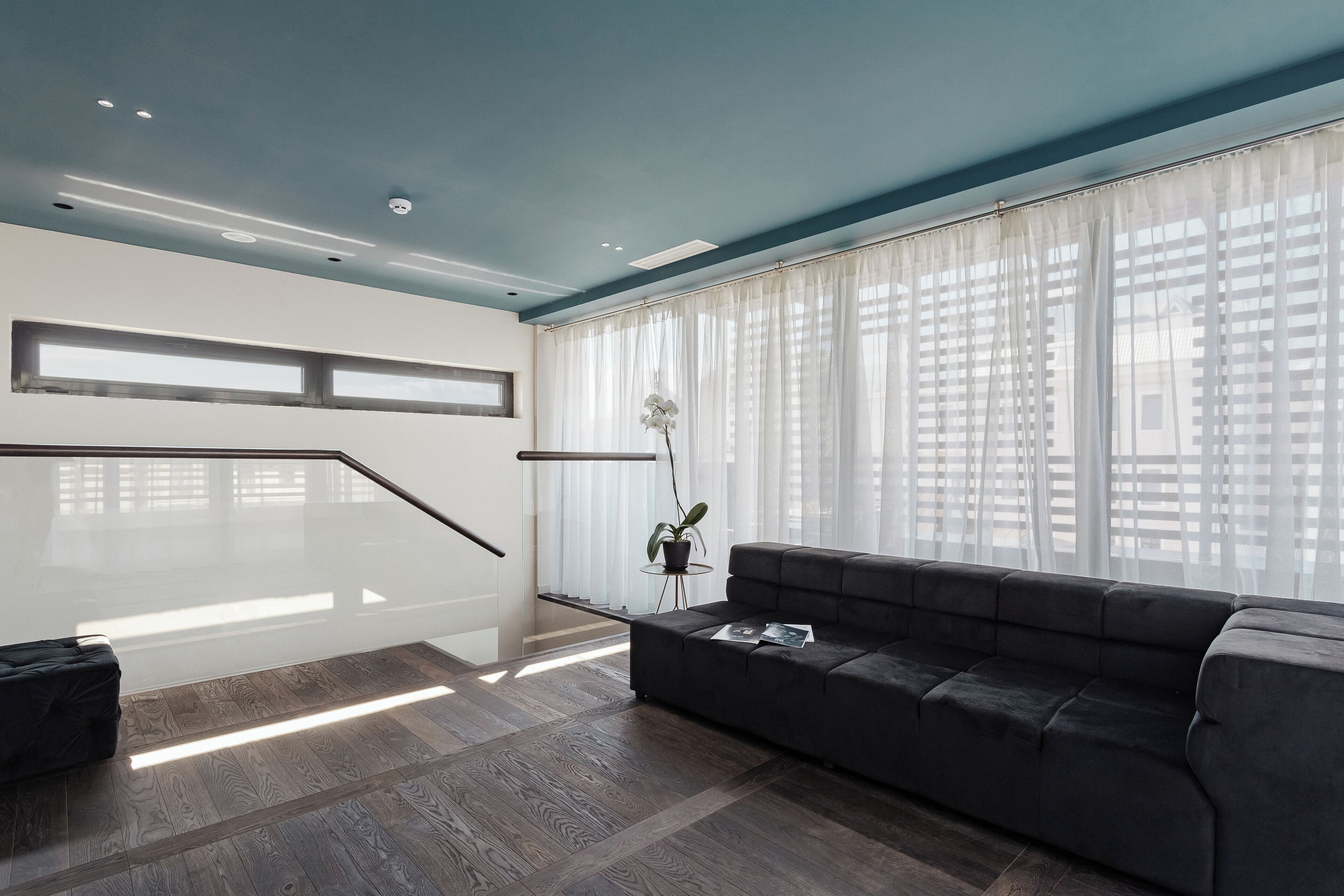 Een moderne woonkamer met een donkergrijze bank, een glazen trap en grote ramen met dunne gordijnen.