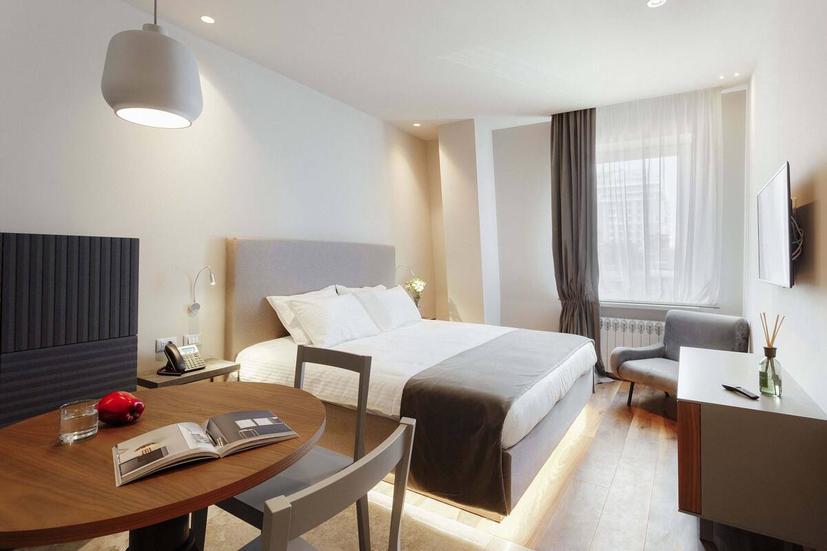 A modern hotel room with a bed, bedside table, lamp, chair, and a desk with a vase.