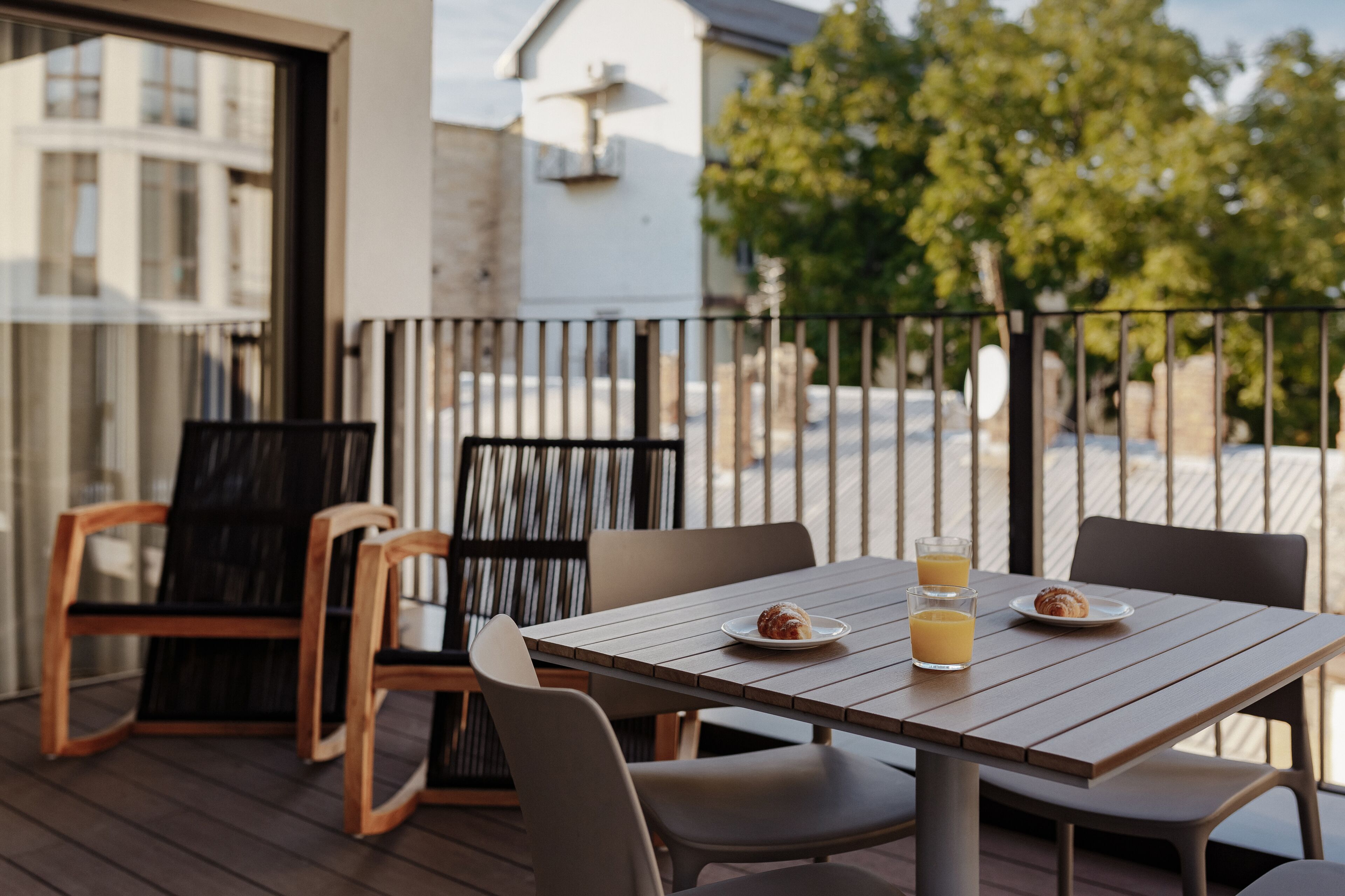 A wooden deck with a table and chairs, two plates with pastries and glasses of orange juice, and a view of a building and trees in the background.