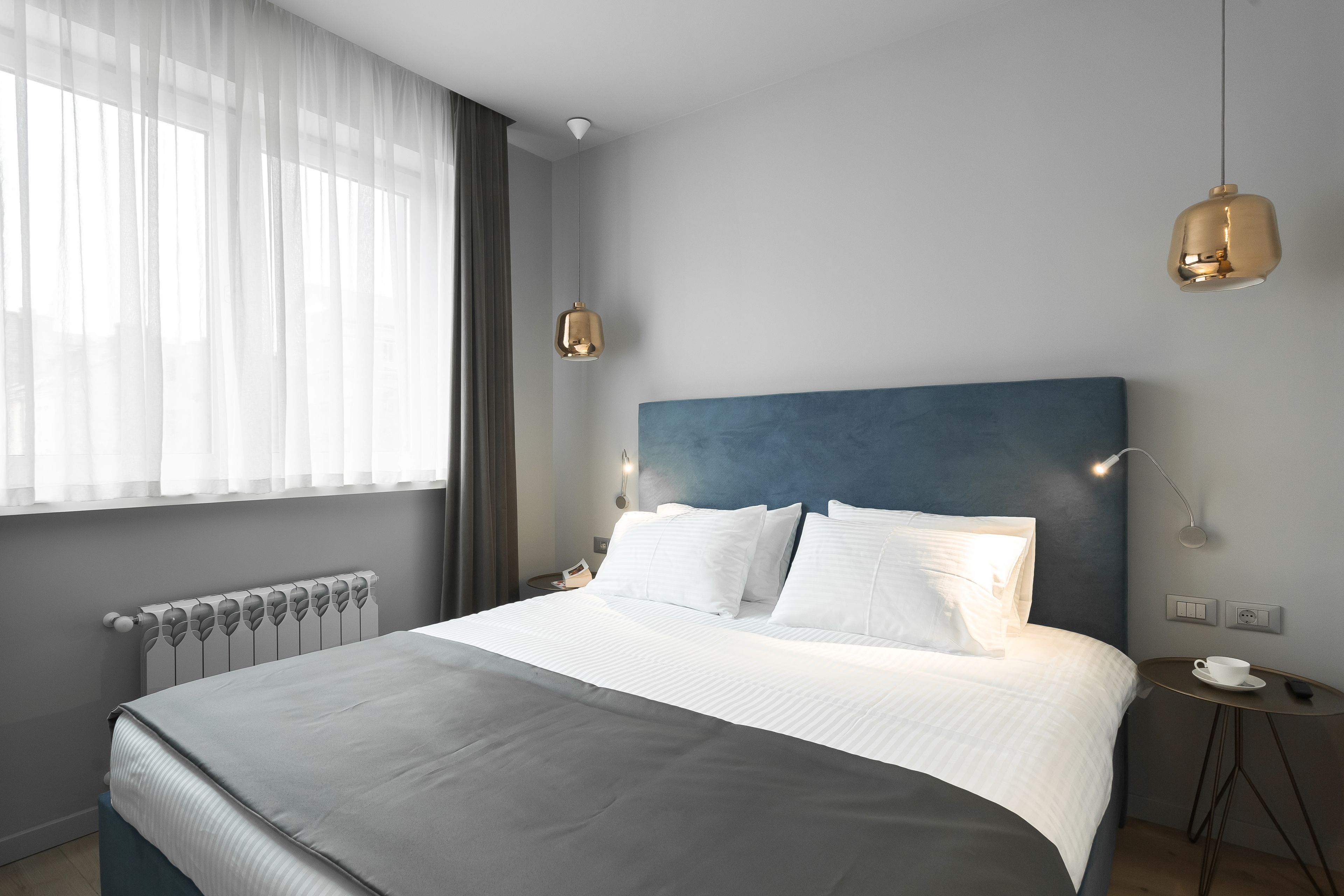 Una habitación de hotel moderna con una cama grande, una mesita de noche, una pequeña mesa redonda y un ventanal con cortinas translúcidas.