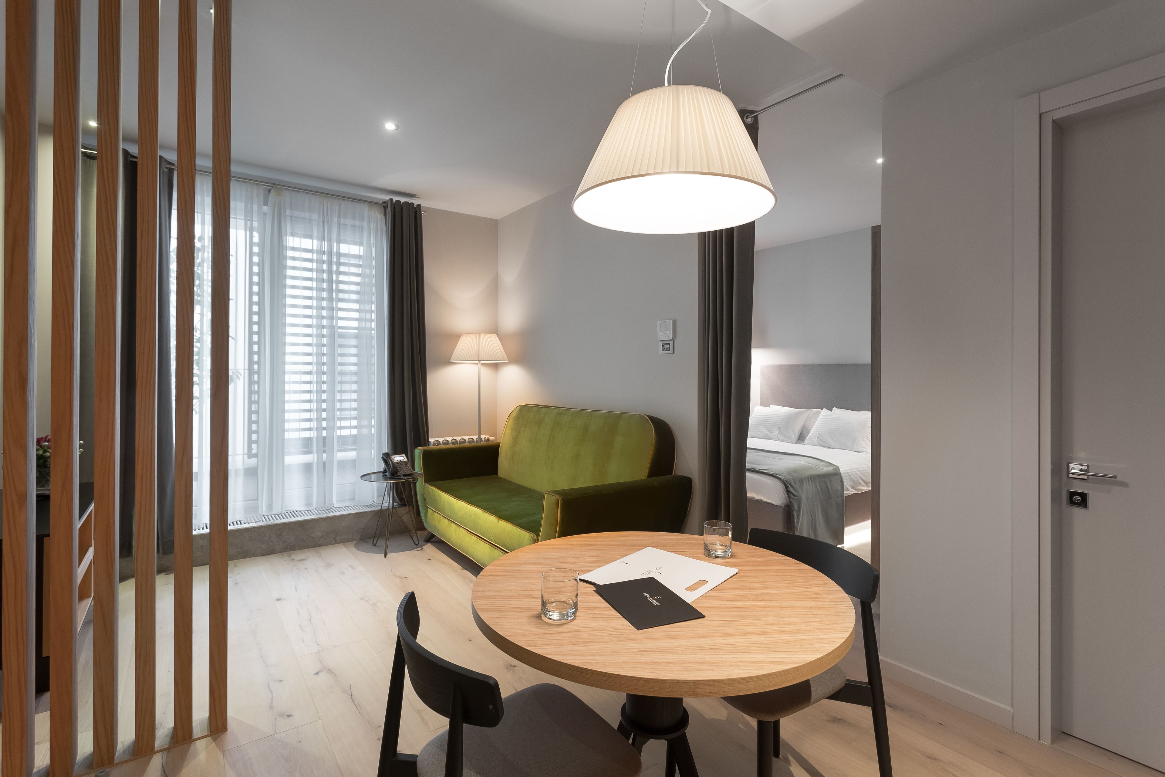 Une chambre d’hôtel moderne avec une table ronde en bois, un canapé vert, une lampe et un lit avec du linge de maison blanc.