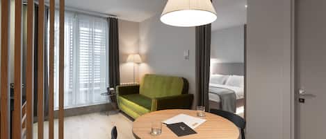 A modern hotel room with a round wooden table, a green sofa, a lamp, and a bed with white linens.