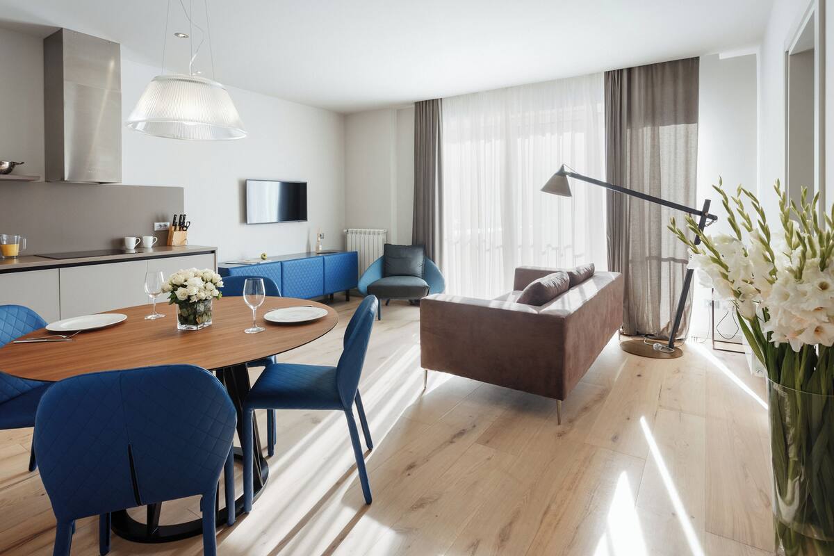 A modern living space with a wooden dining table, blue chairs, a brown sofa, and a large window with curtains.
