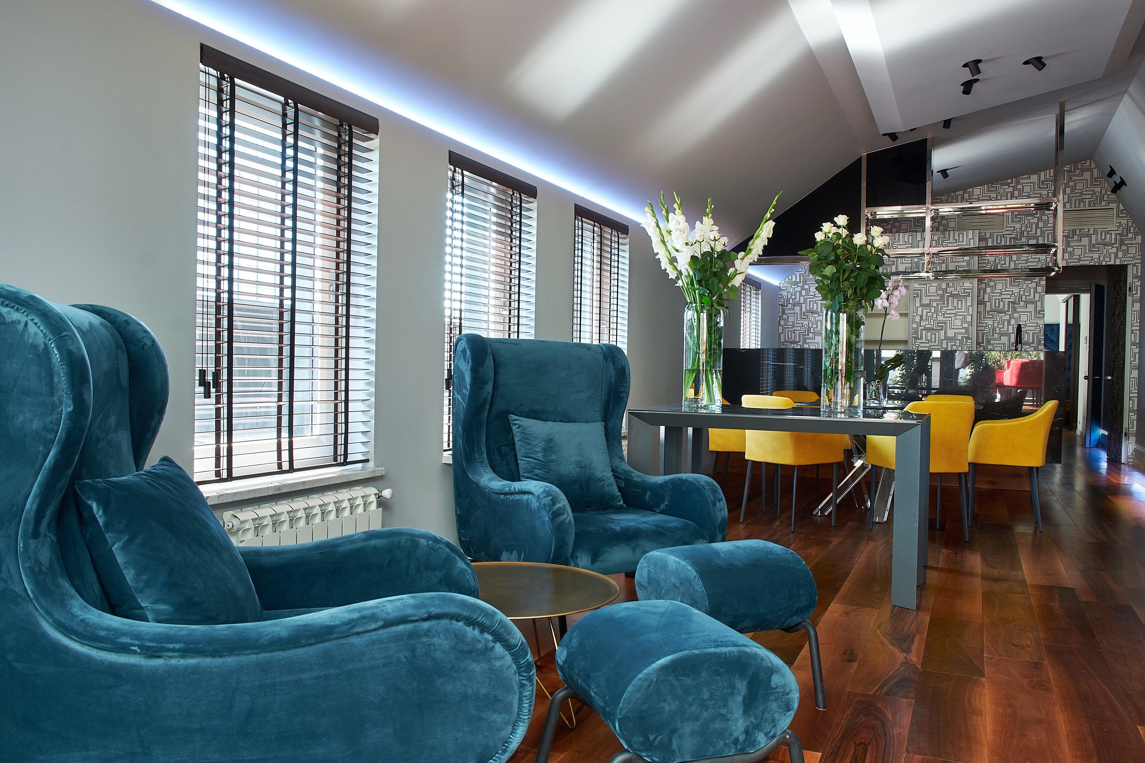 A modern living room with teal sofas, a dining area with yellow chairs, and a kitchen in the background.