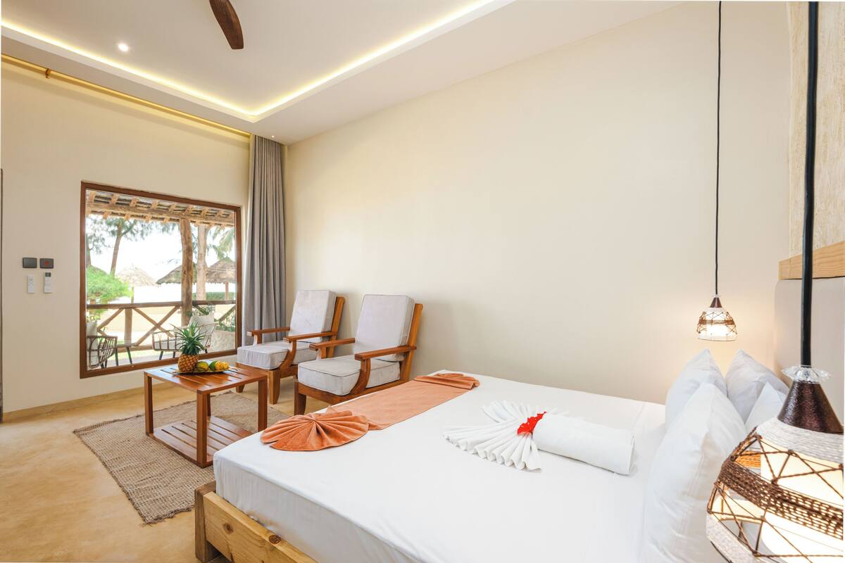 A hotel room with a bed, two chairs, a small table, a window with a view, and a hanging light fixture.