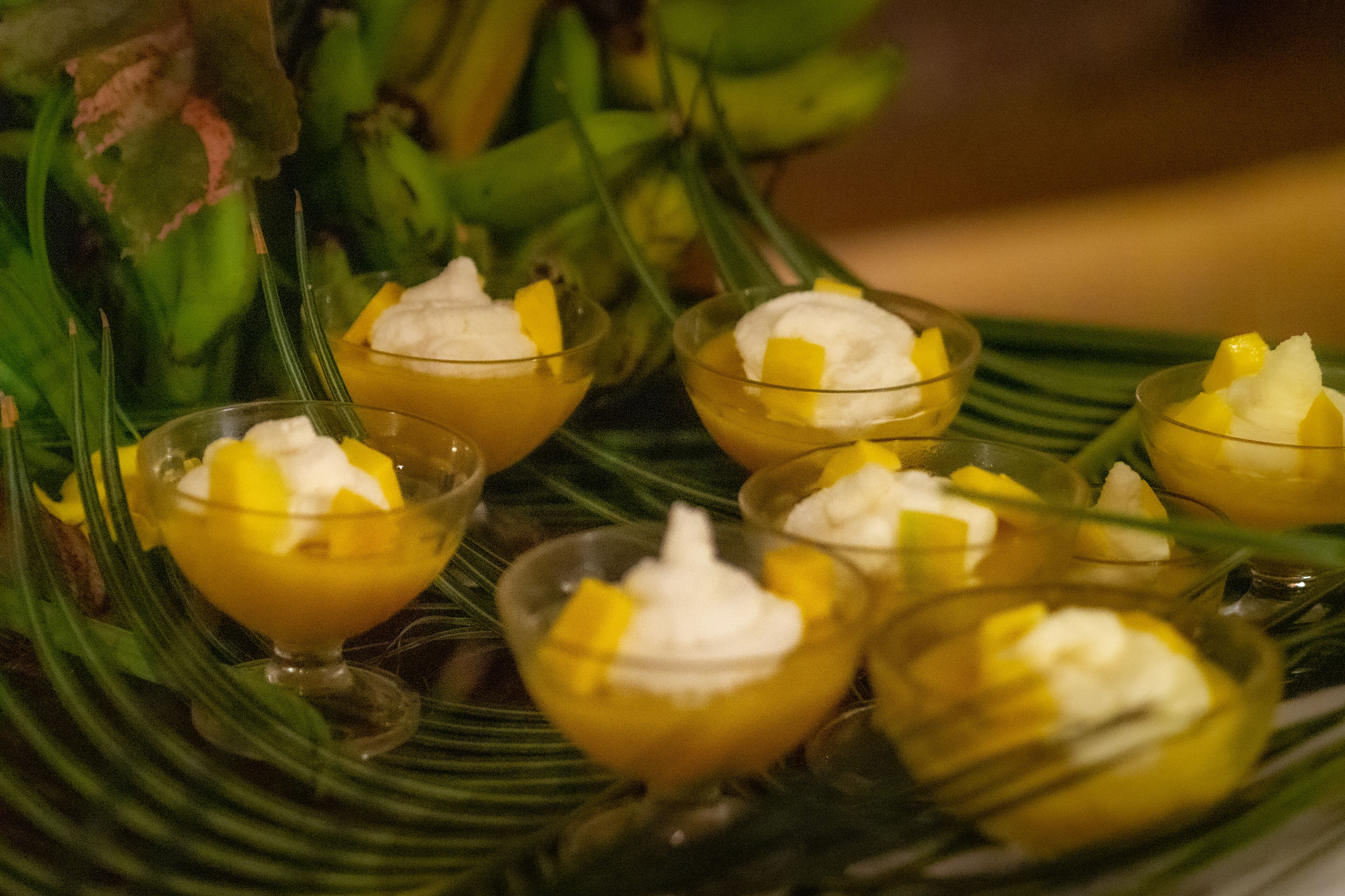 Una bandeja de postres con recipientes pequeños que contienen un postre amarillo cubierto de crema batida y trozos de mango, sobre un plato con estampado de hojas verdes.