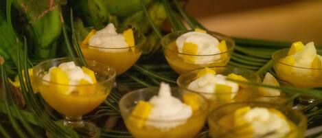 Een etagère met kleine kommetjes gevuld met een geel dessert, afgetopt met slagroom en stukjes mango, geplaatst op een bord met een bladmotief.