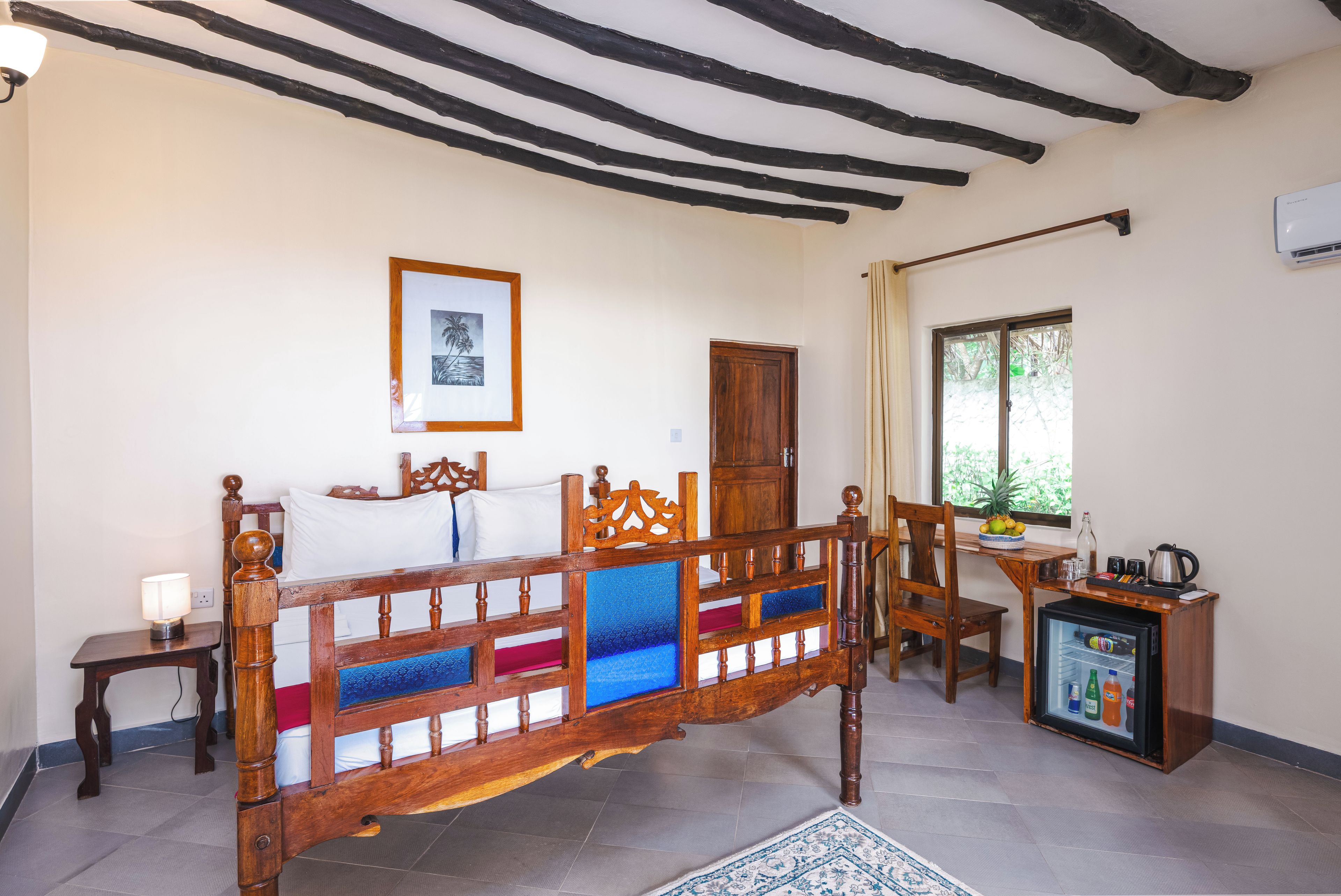 A room with a wooden bed, a small table with a lamp, a refrigerator, and a window with a view of greenery.