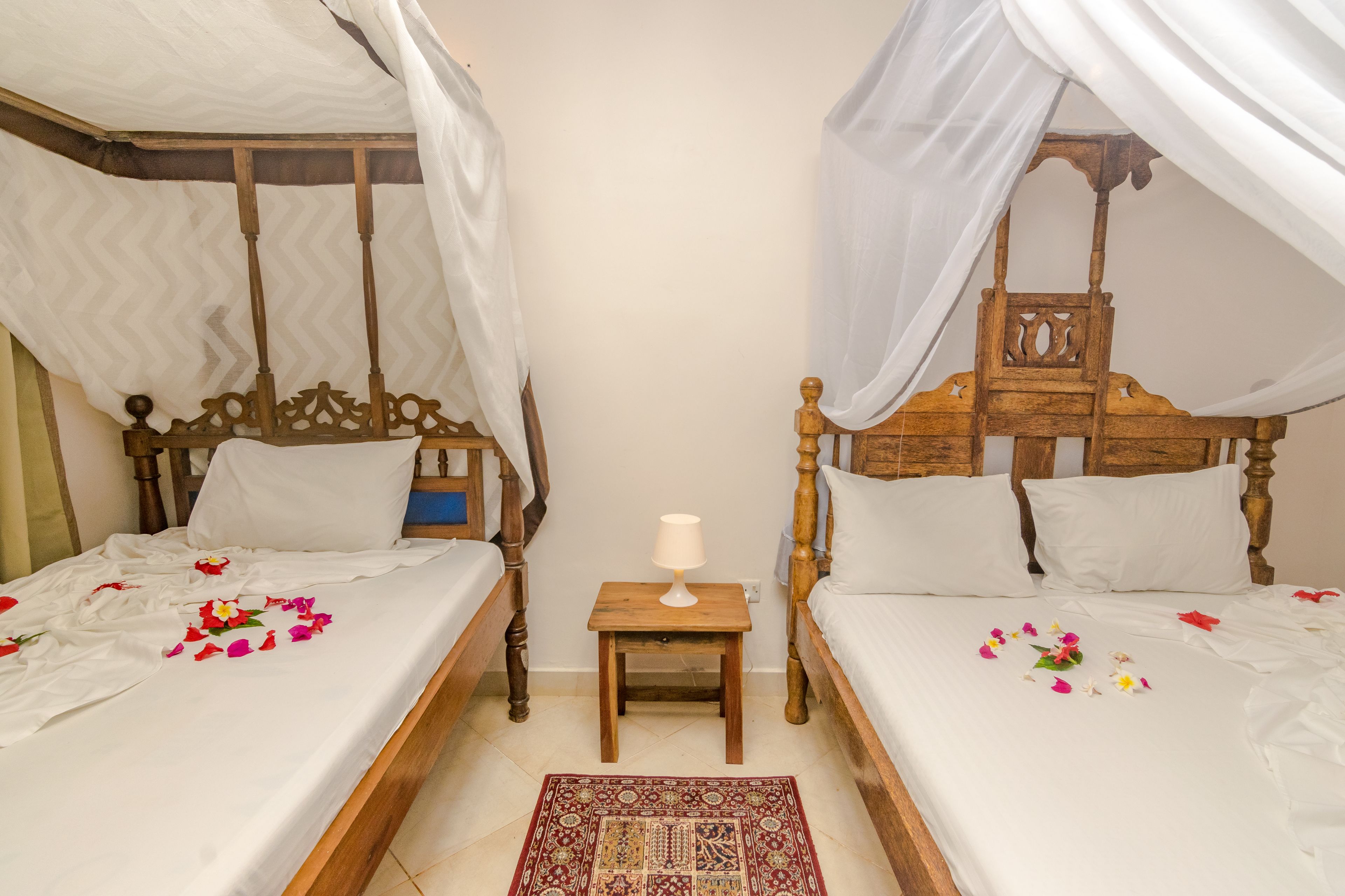 A room with two wooden beds, each with a canopy. There is a small wooden nightstand with a lamp between the beds.