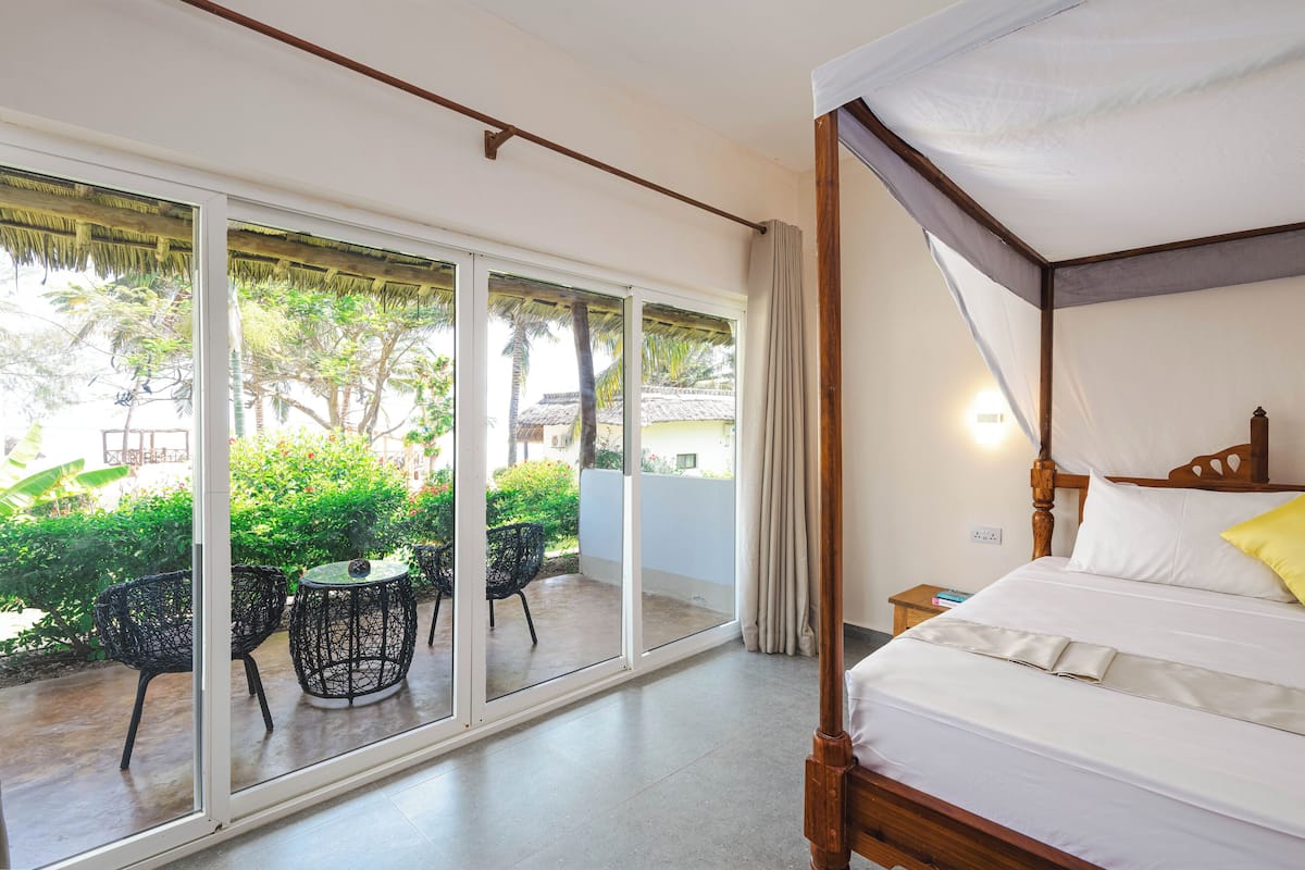 A room with a four-poster bed, a sliding glass door leading to a balcony with outdoor furniture, and a view of greenery and buildings outside.