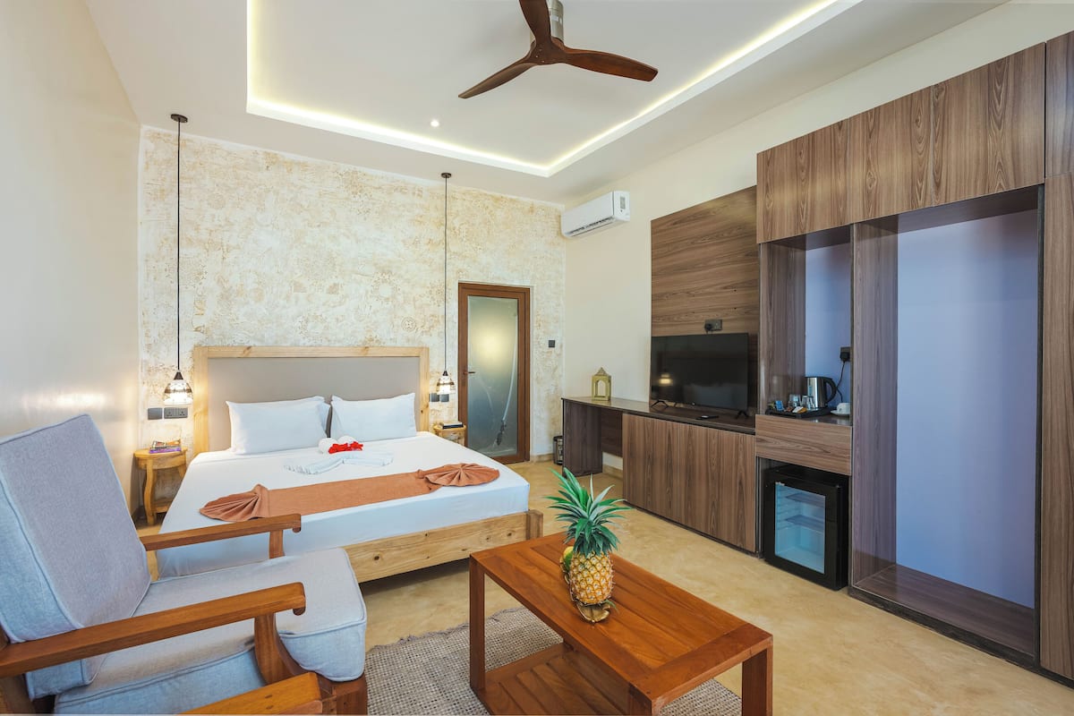 A modern hotel room with a bed, a wooden chair, a small table, a TV, and a minibar.
