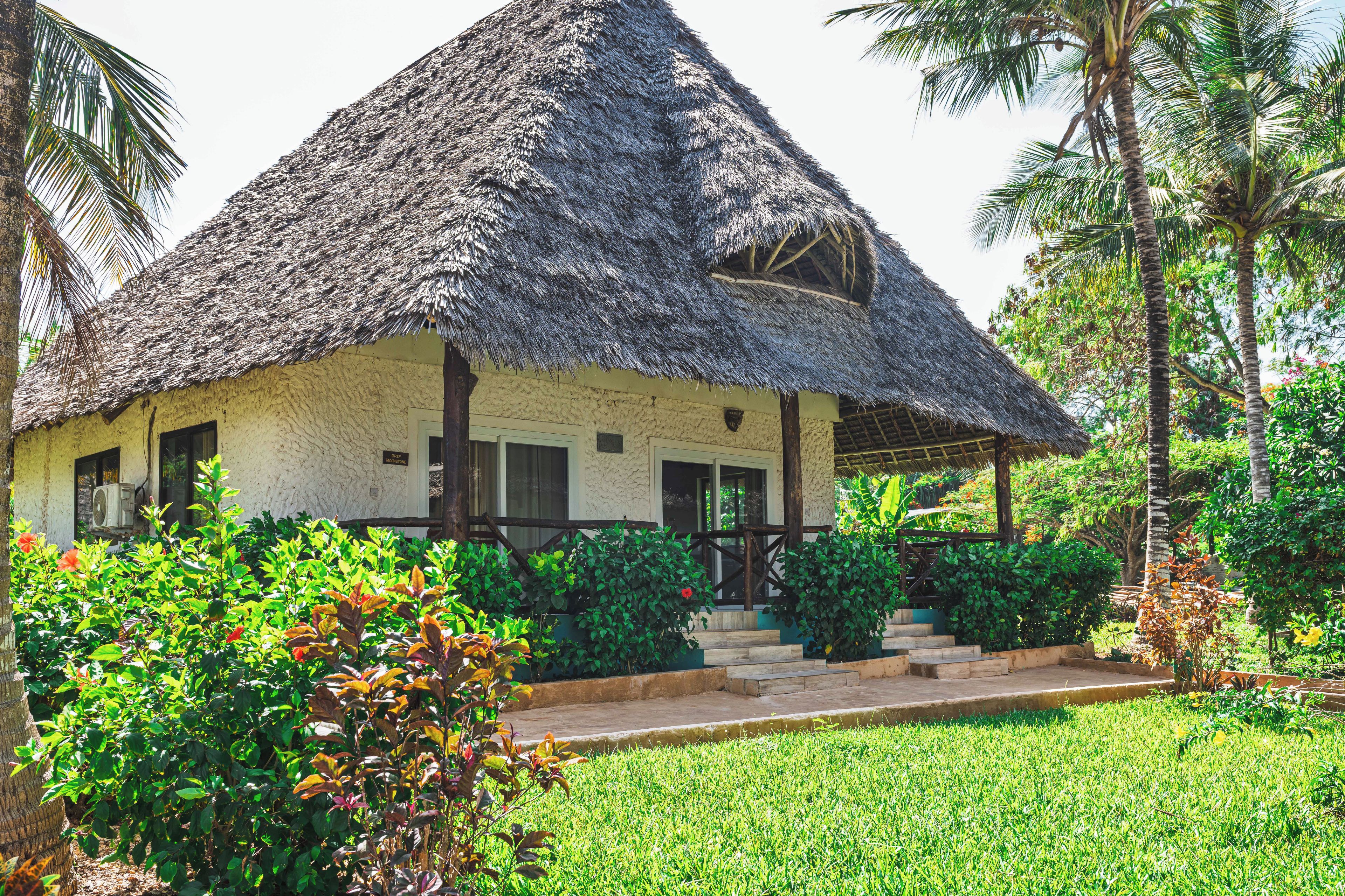 A thatched-roof house with a porch, surrounded by lush greenery and palm trees.