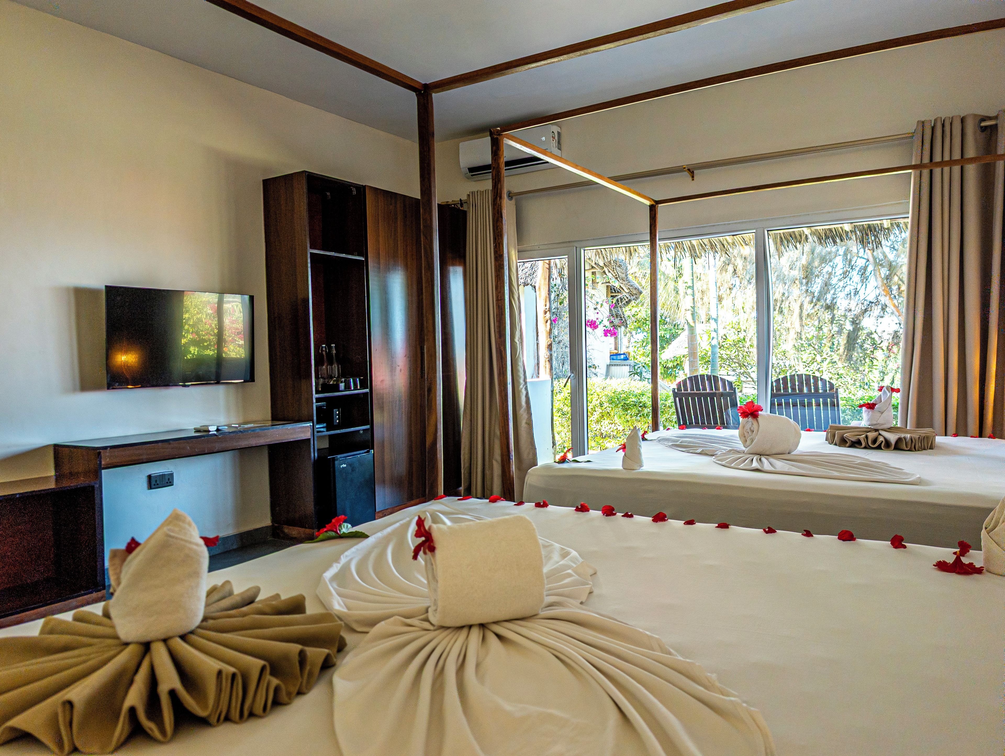 A hotel room with a bed, a TV, a minibar, and a window with a view of greenery.