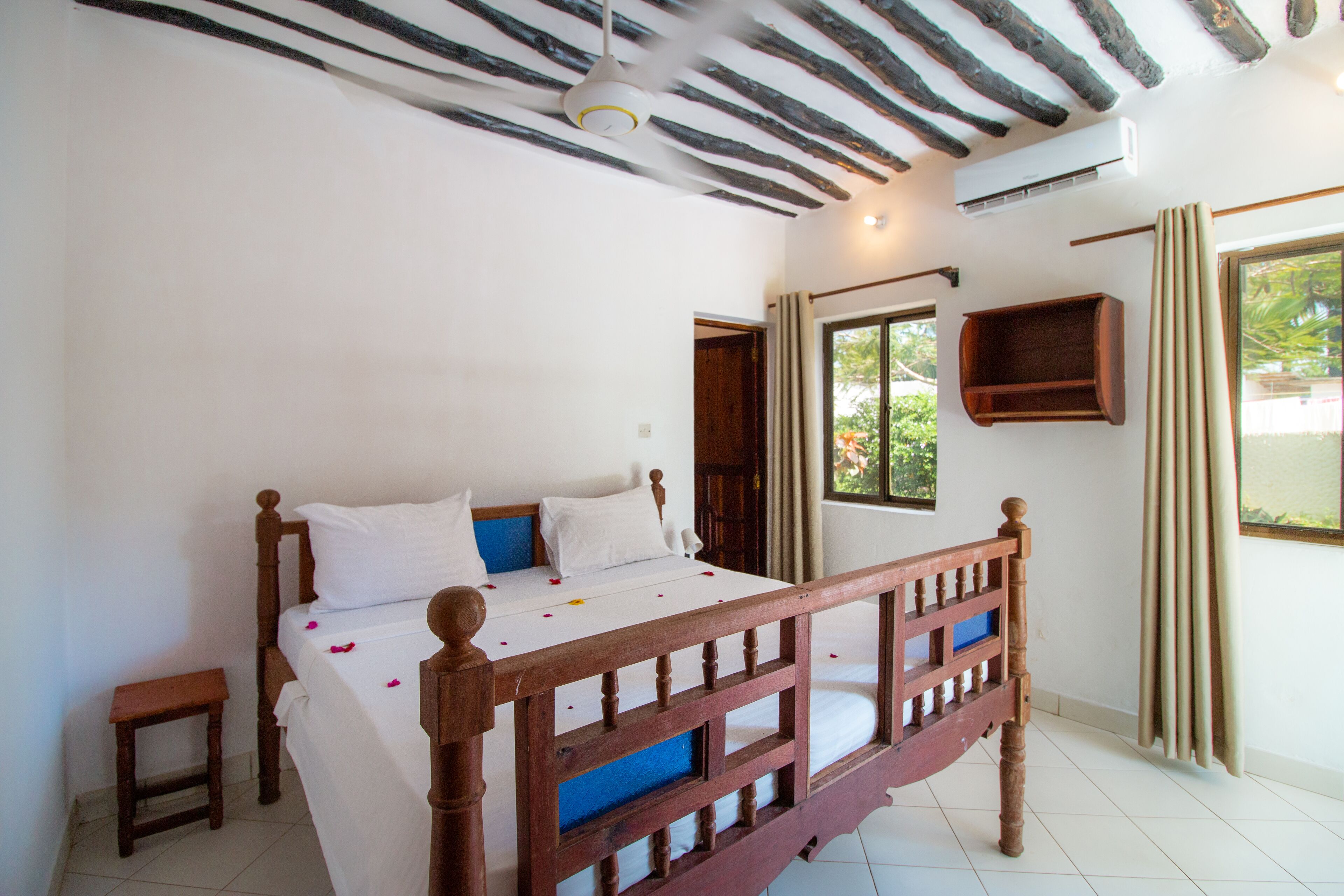 A bedroom with a wooden bed, a small bedside table, a window with curtains, and an air conditioning unit on the wall.