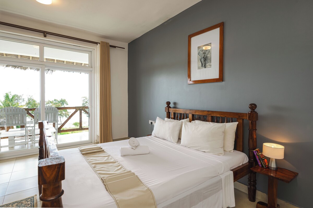 A hotel room with a bed, a nightstand with a lamp, a framed picture on the wall, and a view of the outdoors through a large window.