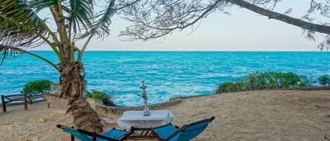 A beach with a table and chairs under a palm tree, overlooking the sea.