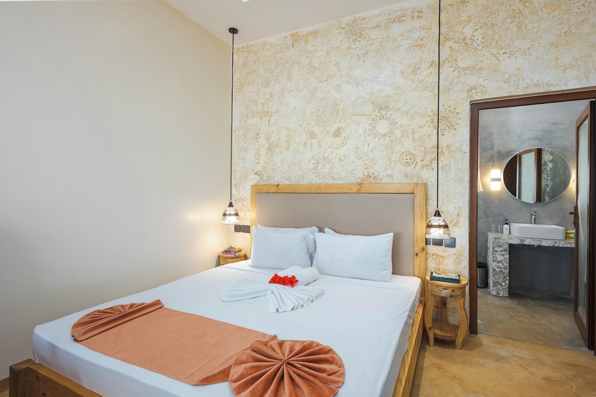 A hotel room with a large bed, a wooden headboard, a bedside table with a lamp, and a bathroom visible through an open door.