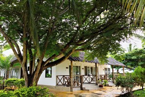 A white building with a thatched roof, surrounded by lush greenery and a large tree. - Tanzanite Beach Resort (Nungwi)