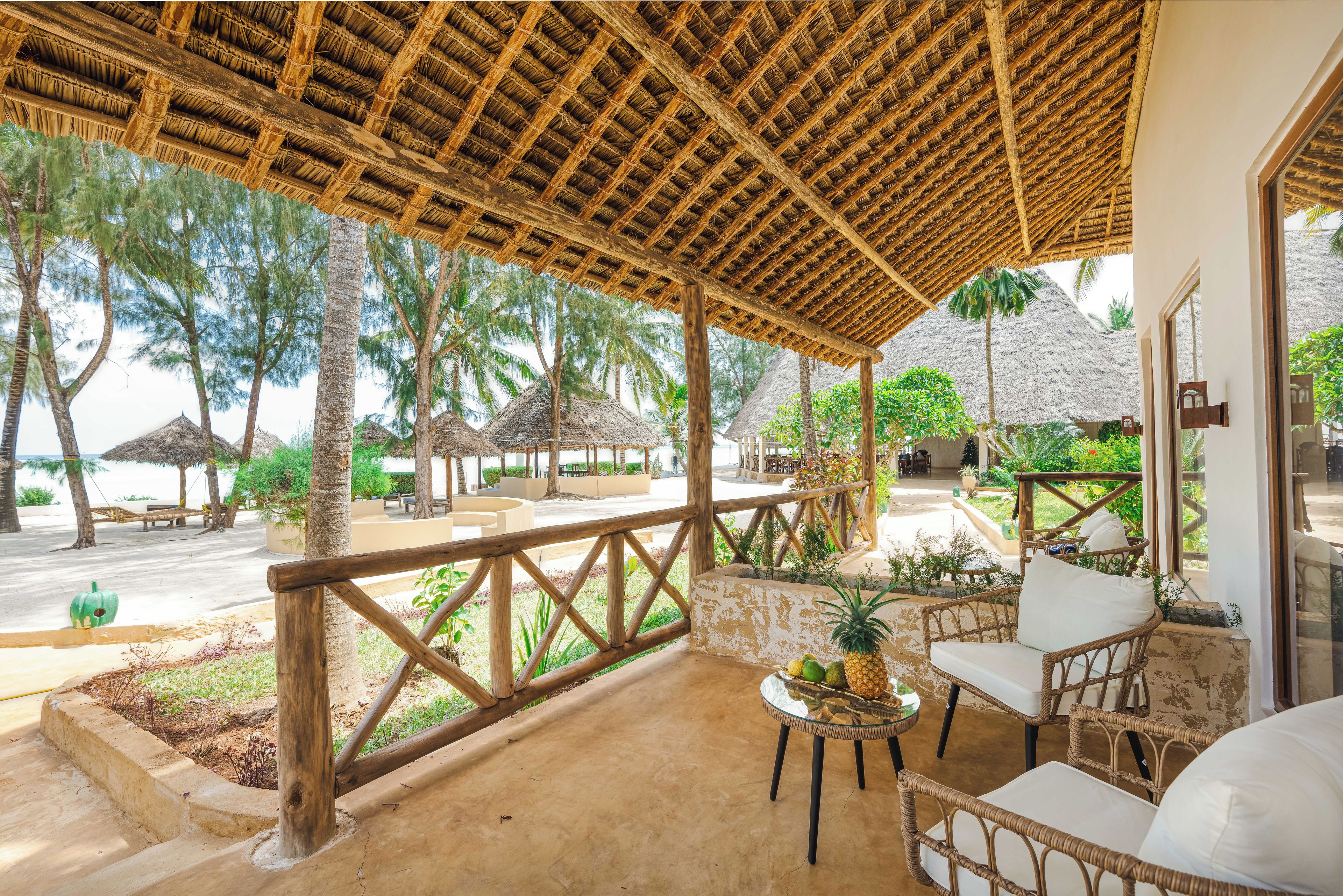 A covered outdoor seating area with wicker furniture, a small table with fruit, and a view of the beach and palm trees.