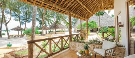 A covered outdoor seating area with wicker furniture, a small table with fruit, and a view of the beach and palm trees.