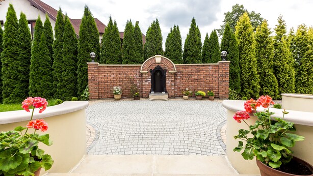 A paved entrance with a brick wall and a black archway, surrounded by potted plants and tall trees. - Hotel Krzyski (Tarnow)