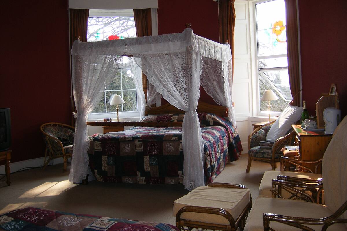 Ein Schlafzimmer mit einem Himmelbett, einer Sitzecke mit Sesseln und einem Fenster mit Vorhängen.