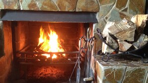 A stone fireplace with a fire burning inside, a metal fireguard, and a stack of firewood to the right. - The Manor Country House Hotel (Dumfries)