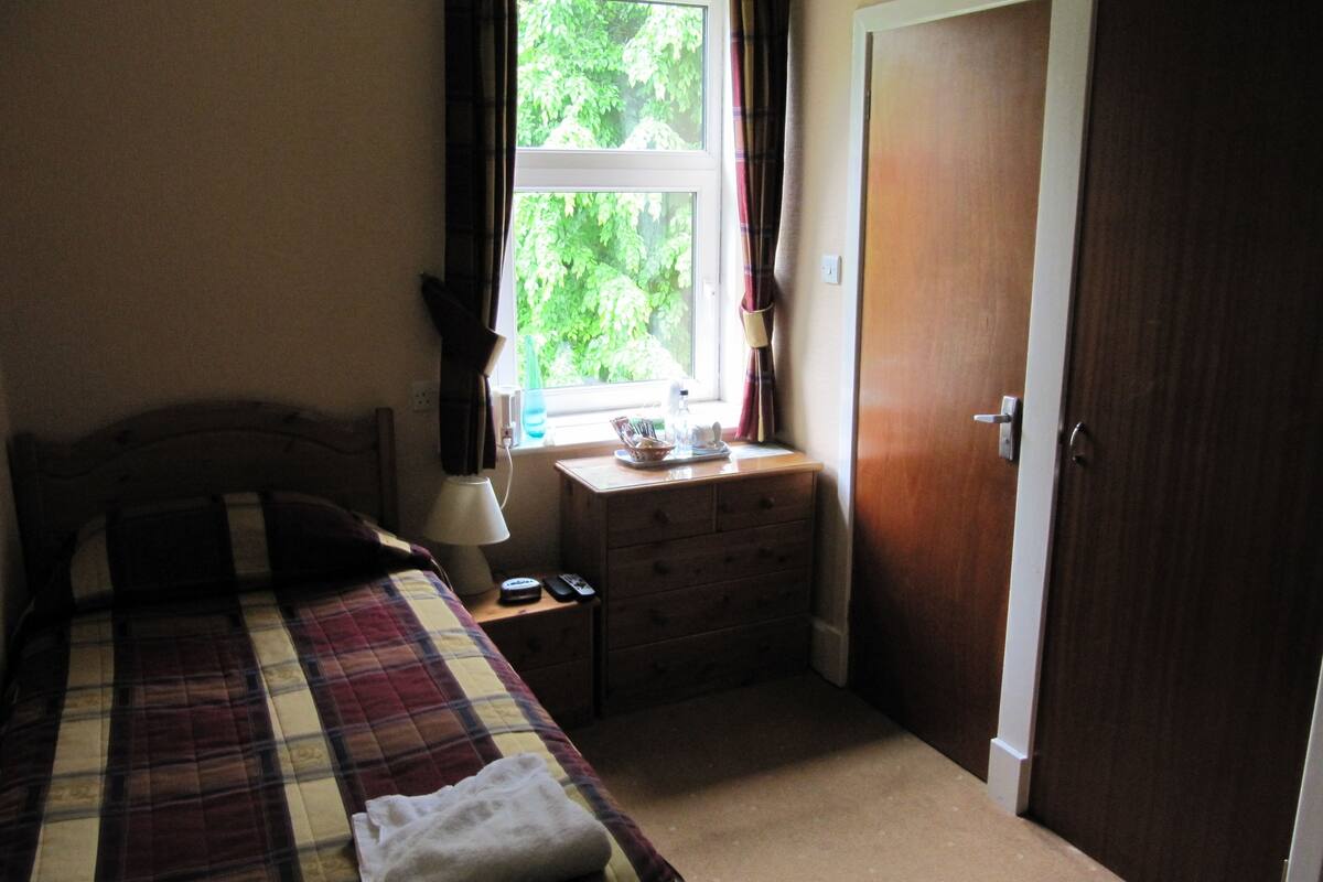 Ein Schlafzimmer mit Bett, Nachttisch, Kommode, einem Fenster mit Vorhängen und einer Tür.