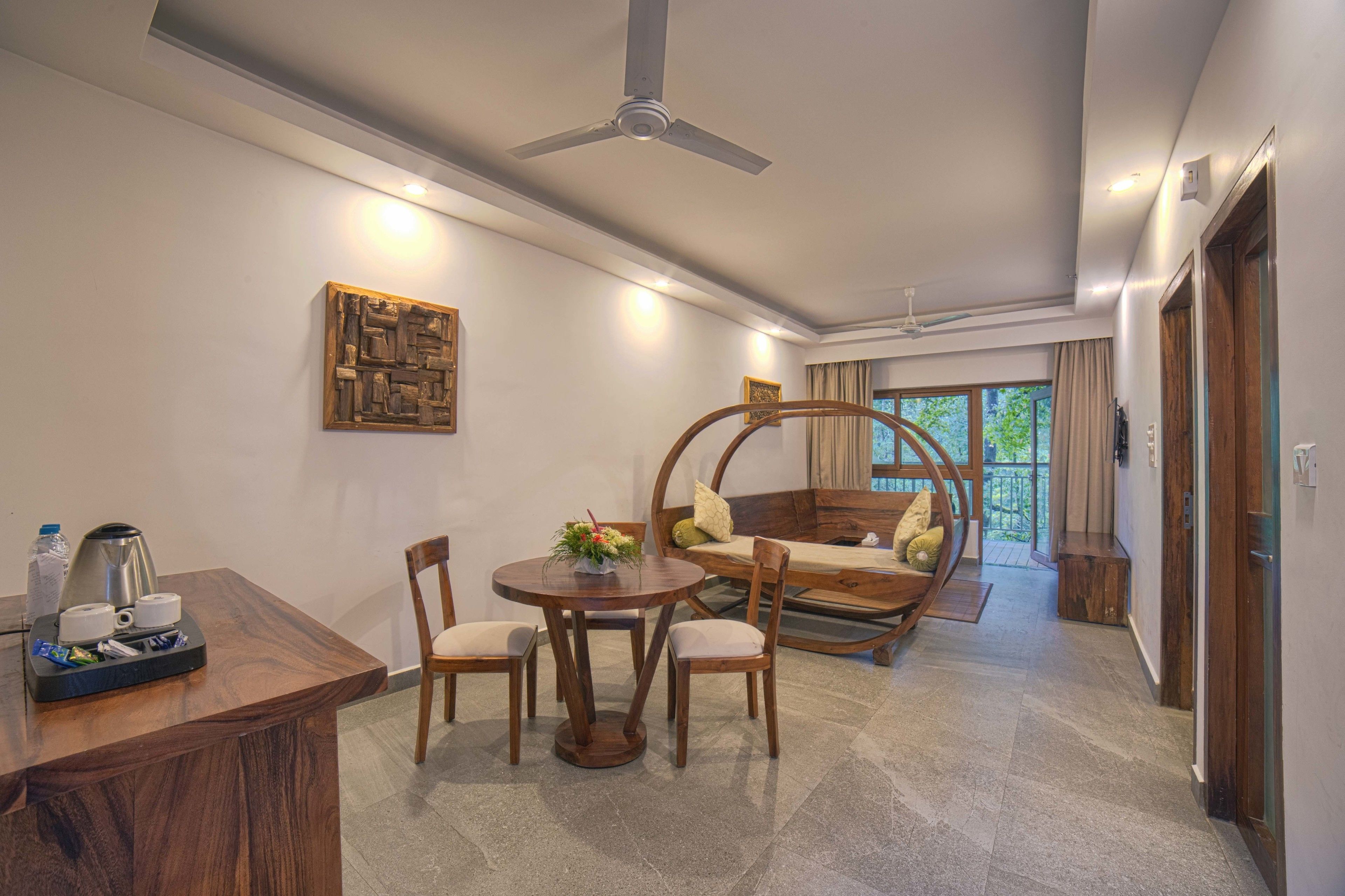 Habitación de hotel moderna con una mesa de comedor de madera, sillas, una mesa redonda de centro con un arreglo floral, una silla colgante tipo hamaca y un ventilador de techo.