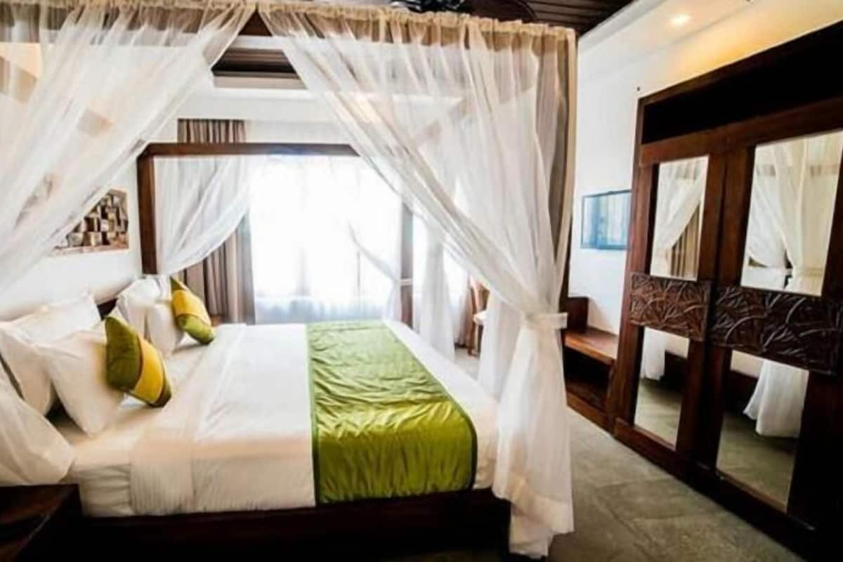 A four-poster canopy bed with white linens and green accents, a large mirror on the wall, and a view of the room's interior.