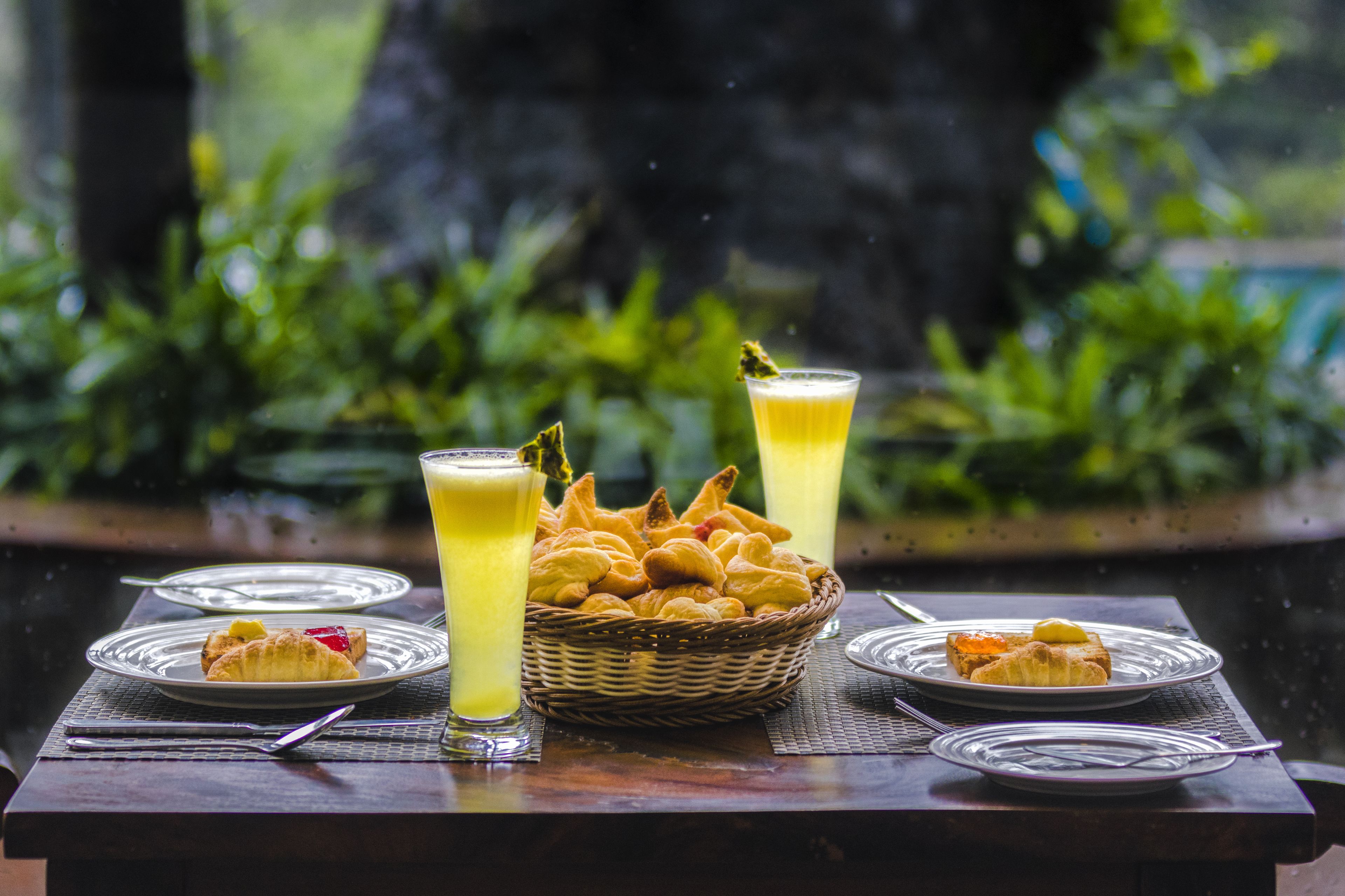A table with plates of food, a basket of pastries, and two glasses of a yellow beverage.