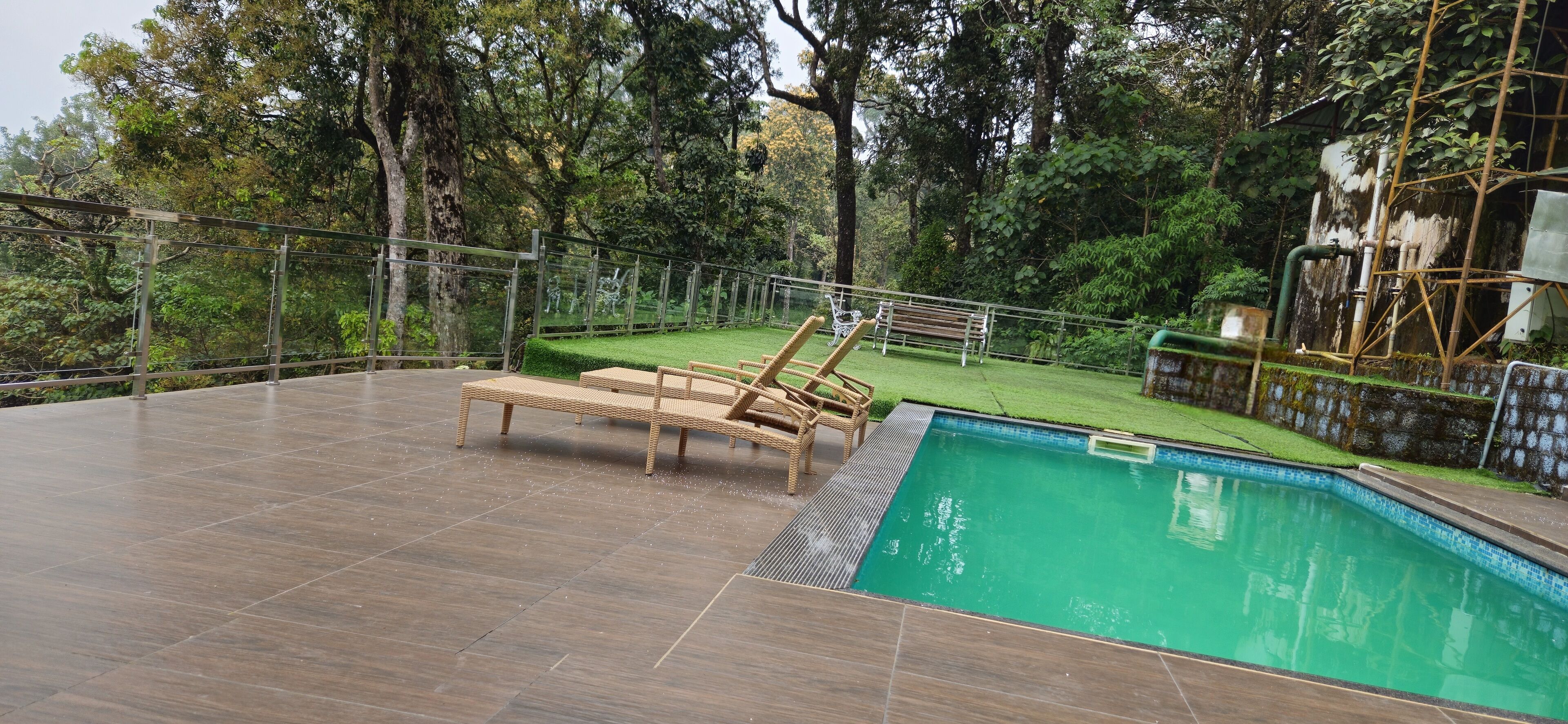 Una piscina con cubierta de madera y sillas de descanso, rodeada por una valla metálica y abundante vegetación.