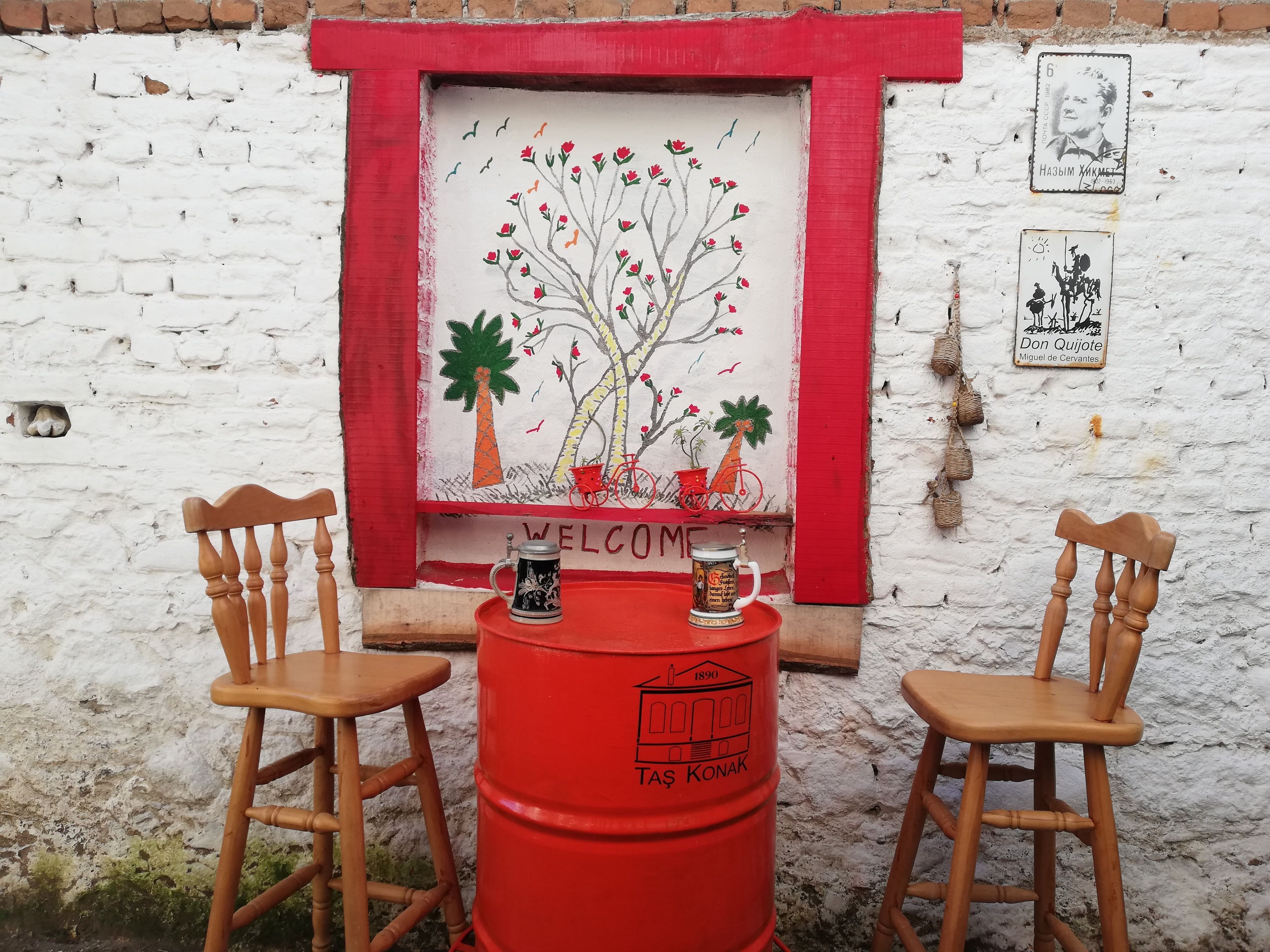 A rustic outdoor seating area with two wooden chairs and a red barrel table, set against a white brick wall with a colorful mural and a welcome sign.