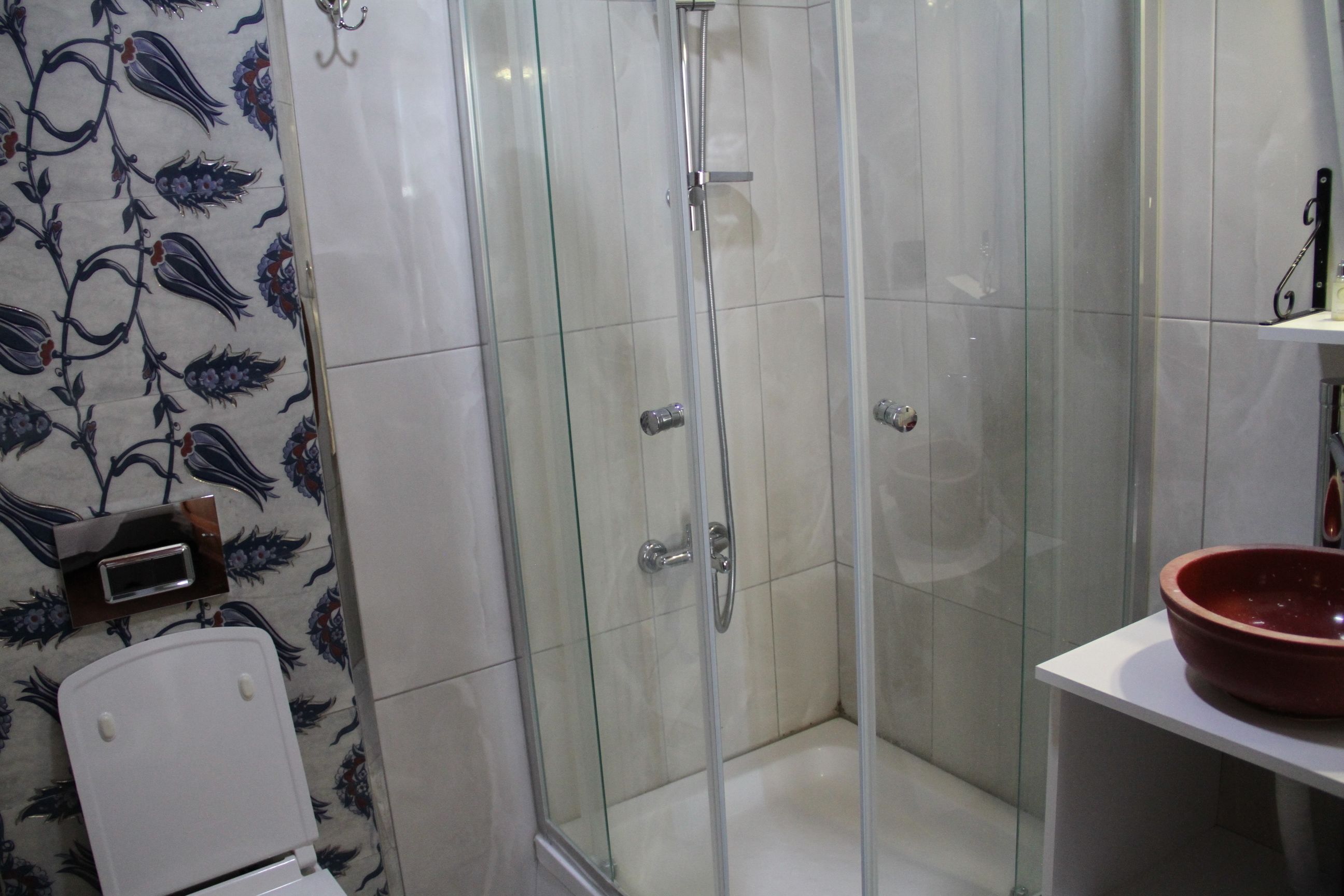 Un baño con cabina de ducha de vidrio, un inodoro blanco y un lavabo rojo sobre un estante blanco.