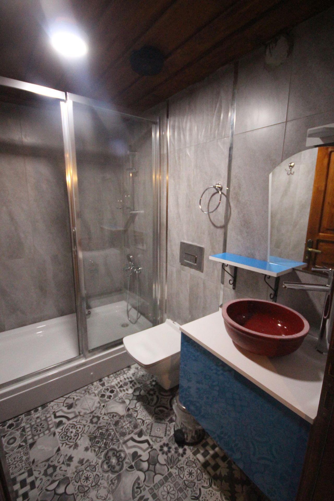 A bathroom with a glass-enclosed shower, a white toilet, a blue vanity with a red bowl sink, and patterned floor tiles.