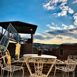 A rooftop terrace with a round table and white metal chairs, overlooking a cityscape. - Bergama Tas Konak (Bergama)