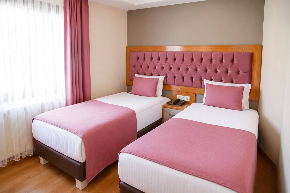 Habitación de hotel con dos camas, cabecero de madera, mesita de noche con teléfono y ventana con cortinas.