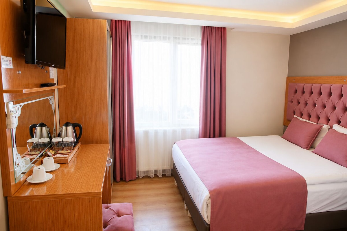 Ein Hotelzimmer mit Bett, Schreibtisch mit Kaffeemaschinen, Fernseher und einem Fenster mit Vorhängen.