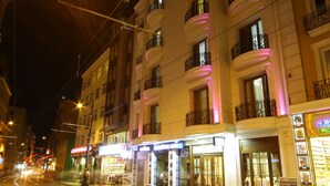 A street with tram tracks, illuminated buildings with balconies, and storefronts.