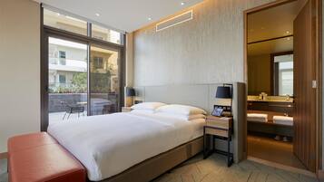 A modern hotel room with a large bed, a bedside table with a lamp, a red bench, a sliding glass door to an outdoor area, and a wooden wardrobe with a mirror.