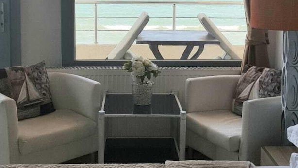 Ein Hotelzimmer mit großem Fenster und Blick aufs Meer, einem Bett, zwei Sesseln, einem kleinen Tisch mit einer Vase und einer Lampe.