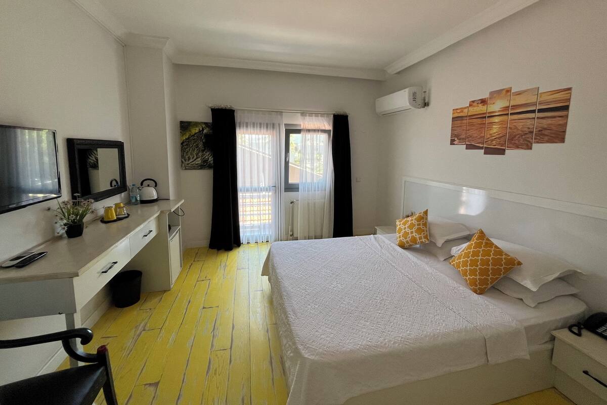 Ett hotellrum med en säng, ett skrivbord med en stol, en tv, ett fönster med gardiner och konstverk på väggen.