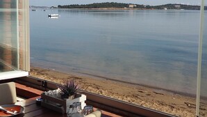 Blick aus einem Fenster auf ruhiges Wasser mit Booten, einen Sandstrand und Gebäude in der Ferne.