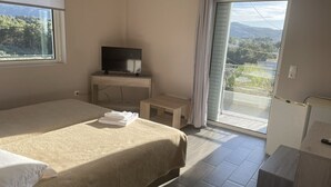 A hotel room with a bed, a TV on a stand, a small table, and a balcony with a view of the outdoors. - Hotel Paradise (Oropos)